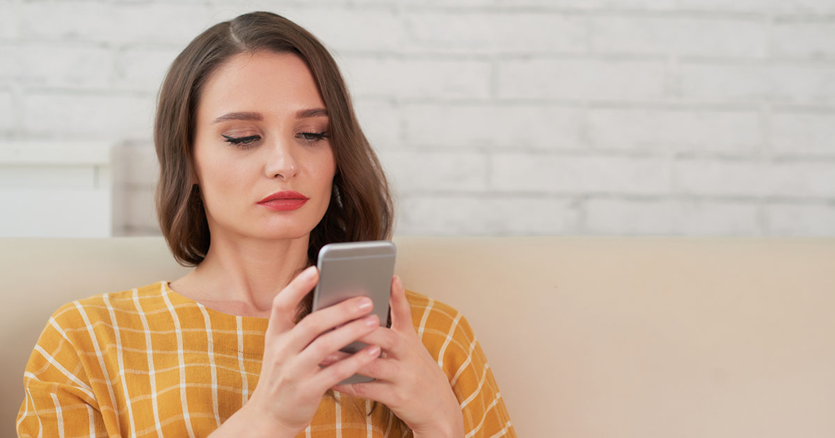 Nő ül a kanapén és a telefonján olvas, a figyelme elfelé irányul, egy idegen férfival beszélget – a mikromegcsalás és az online intimitás eltolódásának vizuális metaforája