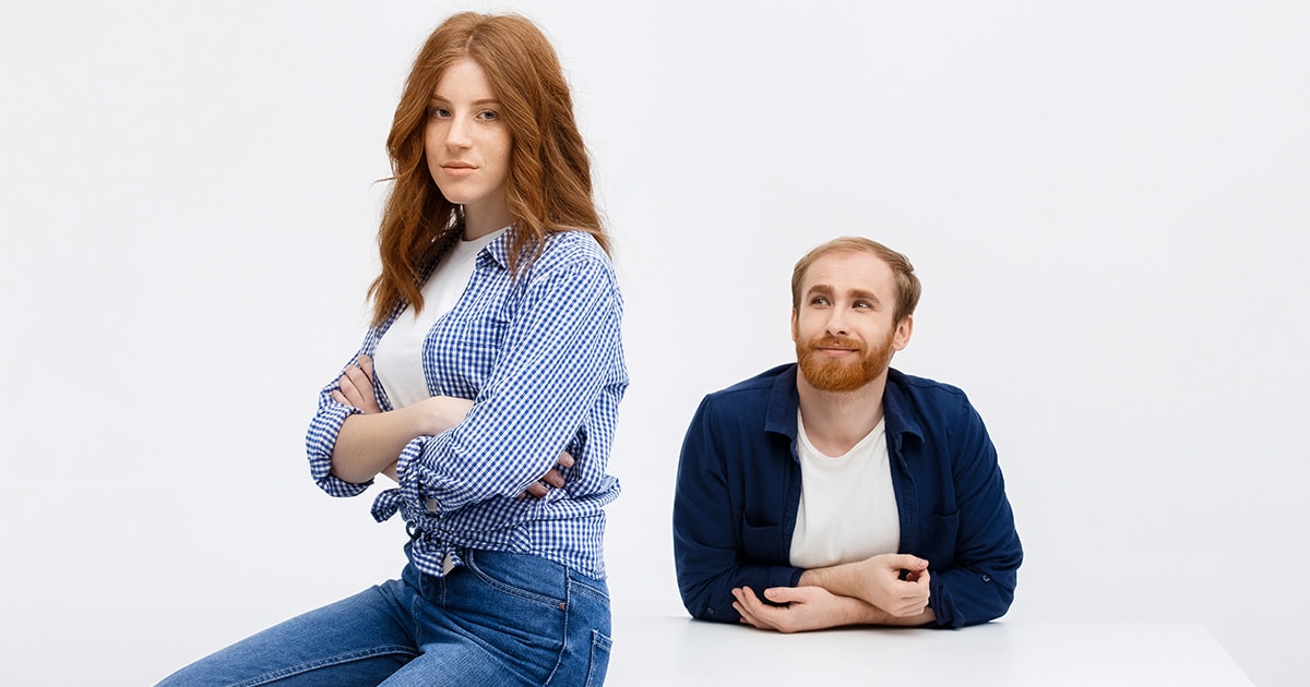 Miért választják egyre többen az egyedüllétet a párkapcsolat helyett? Egy nő komolyan ül az asztal szélén karba tett kézzel, miközben mögötte egy férfi boldogan néz rá, de a nő elérhetetlen, mert nem akar kapcsolatot senkivel.