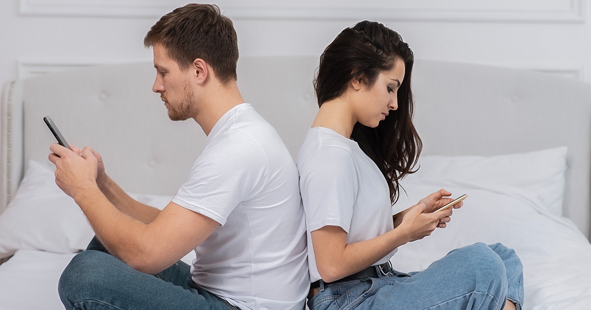 Telefonfüggőség a párkapcsolatban – a valódi kapcsolódás ára Fiatal pár egymásnak háttal ül az ágyon, mindketten a telefonjukat nézik, érzelmi távolság van köztük.